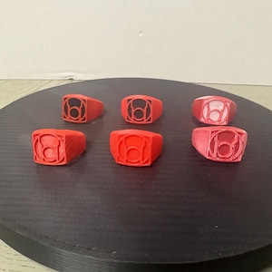 Puede incluir: Seis anillos rojos dispuestos en dos filas, cada uno con un emblema circular en relieve con un símbolo central que se asemeja a una linterna. Los anillos tienen un acabado mate y un diseño de banda gruesa, con uno mostrando una incrustación blanca.