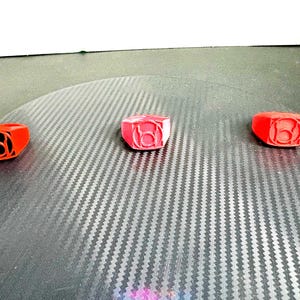 Puede incluir: Tres anillos rojos con un diseño gráfico negro se muestran sobre una superficie texturizada. Los anillos están espaciados uniformemente y parecen estar hechos de plástico o un material similar. El diseño de los anillos es un símbolo estilizado.