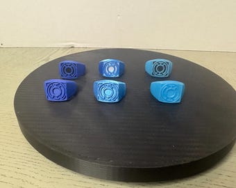 May include: Six rings in various shades of blue, each featuring a circular emblem with a central dot and surrounding lines. The rings are arranged in two rows of three, displaying a range of blue tones from dark navy to a lighter sky blue.