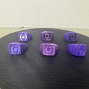Puede incluir: Seis anillos de color morado, cada uno con una cara cuadrada y un emblema circular en el centro. Los anillos muestran variaciones en la intensidad del color y el acabado, con un anillo que presenta un centro circular blanco y otros con diseños morados sólidos.
