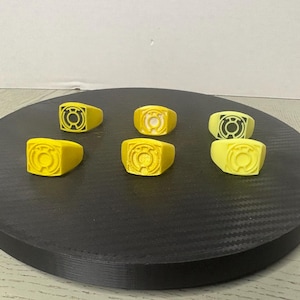 May include: Six yellow rings arranged in two rows, each featuring a raised circular emblem with a central dot and surrounding lines. The rings have a smooth, matte finish and a square-shaped face, displaying a consistent design across all pieces.