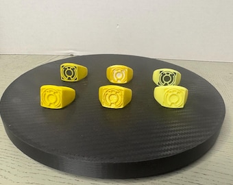 May include: Six yellow rings arranged in two rows, each featuring a raised circular emblem with a central dot and surrounding lines. The rings have a smooth, matte finish and a square-shaped face, displaying a consistent design across all pieces.