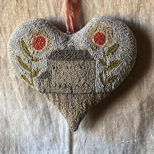 Op de afbeelding: Een hartvormig naaldviltkussen met een bruin en beige huisontwerp en rode en gele bloemen. Het kussen hangt aan een rood lint.