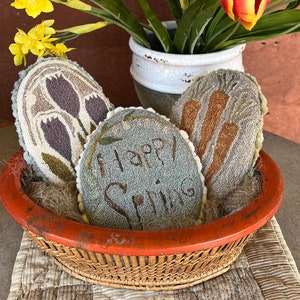 Op de afbeelding: Een rieten mand gevuld met drie naaldviltkussens. De kussens zijn versierd met lentethema's, waaronder tulpen, wortels en de woorden "Happy Spring". De mand staat op een tafel met een bloemstuk van gele narcissen en oranje tulpen in een witte vaas.