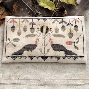May include: A rectangular fabric piece featuring a cross-stitch design on a light beige background. The embroidery depicts two dark brown turkeys facing a central floral motif, surrounded by hanging acorns and leaves in muted earth tones.