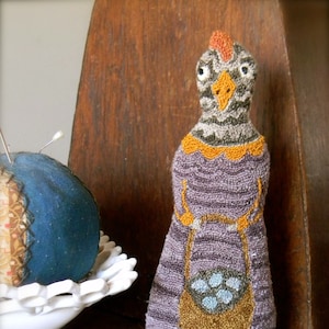 Op de afbeelding: Een gebreide pop in paars en bruin met een grijs en oranje hoofd, die een bruine tas met blauwe bloemen vasthoudt. De pop staat op een houten oppervlak.