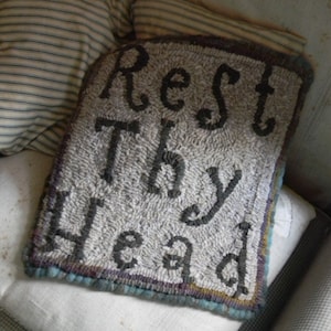 May include: A rectangular, gray, textured pillow with the words "Rest Thy Head" embroidered in dark gray. The pillow has a dark brown border with hints of blue and purple. It rests on a white pillow and a patterned cushion.