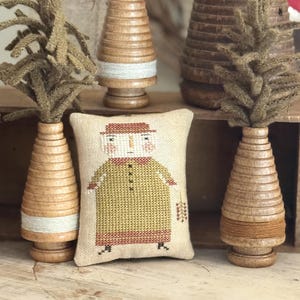 May include: A rectangular fabric pincushion featuring a cross-stitched figure in a yellow dress and red hat, flanked by three wooden spools topped with olive green felt sprigs, arranged on a light-colored wooden surface.