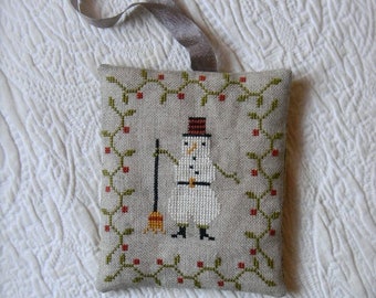 May include: A hand-stitched ornament featuring a snowman holding a broom, surrounded by a green and red holly border.
