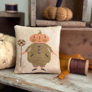 May include: A cross-stitched pillow featuring a pumpkin-headed figure in a green outfit, holding a flower-like object. The pillow is on a wooden surface with a spool of brown thread and a small, felt pumpkin in the background. The pillow measures approximately 5 inches square.
