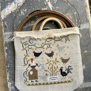 May include: A beige linen bag with a cross-stitch design of a woman and chickens in a garden. The bag is hanging from a wire hoop and has a brown and white checkered fabric lining.