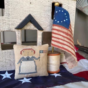 May include: A small, beige pillow with a cross-stitch design of a woman holding a miniature American flag. The pillow is sitting on a red, white, and blue fabric with stars.