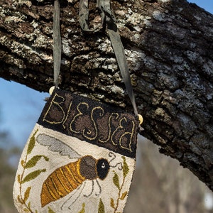 May include: A small, hand-stitched bag with a bee design, hanging from a tree branch. The bag features the word "BLESSED" embroidered across the top. The bee is yellow and brown, with green leaves around it. The bag has a dark grey strap.