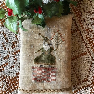 May include: A cross-stitch pillow featuring a green and brown figure standing on a red and white checkered platform. The figure is holding a white object with red accents. The pillow is surrounded by a brown border and is sitting on a white lace tablecloth.