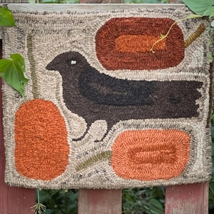 May include: Hand-hooked wall hanging featuring a black crow and three orange pumpkins on a beige background. The crow is centered, with pumpkins on either side and above. The piece is framed with a neutral border and is mounted on a red wooden fence.