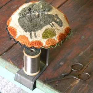May include: A brown and orange needlepoint pincushion with a green and gray sheep design. The pincushion is attached to a metal stand with a spool of thread.
