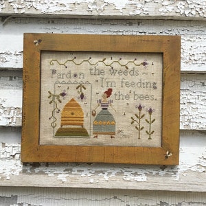 May include: A framed cross-stitch with a beekeeper and a beehive. The text reads "Pardon the weeds... I'm feeding the bees."