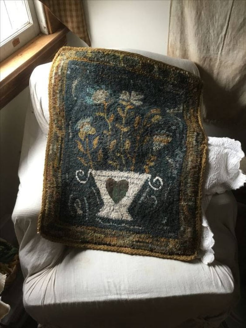 May include: A hand-stitched rug with a floral design in shades of blue, green, and yellow. The rug features a vase with a heart in the center, surrounded by stylized flowers. The rug is framed with a gold border.