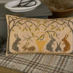 May include: A beige pillow with a cross-stitch design of a tree with five rabbits underneath. The rabbits are in various colors, including brown, gray, and white. The tree has green leaves and pink flowers.