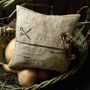May include: A beige needlepoint pillow with the text "Eliza May's Needlework" embroidered in black thread. The pillow is sitting in a wicker basket with dried flowers and a feather.