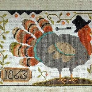 May include: Cross-stitch embroidery of a gray turkey wearing a black top hat with a key above its head. The turkey has orange and brown feathers and is standing on a brown surface. The year 1863 is stitched in a brown rectangle below the turkey.