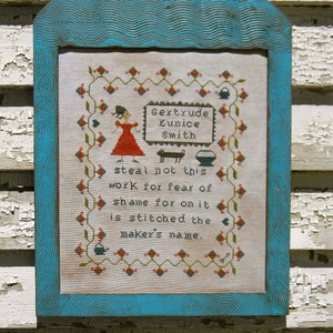 May include: A framed cross-stitch sampler with a blue frame and a white background. The sampler features a floral design with a teapot, a cat, and a woman in a red dress. The text reads "Bextrude Eunice Smith steal not this work for fear of shame for on it is stitched the maker's name."