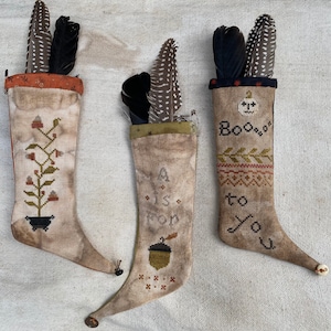 May include: Three handmade stockings with a pointed toe and a button at the end. The stockings are decorated with different embroidery designs. The first stocking has a green plant with red berries and a black pot. The second stocking has a brown acorn and the text "A is for on". The third stocking has a white pumpkin face and the text "Boo to you".