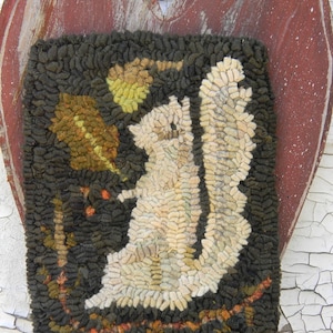 May include: A hand-stitched rug depicting a white squirrel with a bushy tail, sitting on a branch with green and brown leaves. The rug is framed by a dark green border and is mounted on a red wooden heart.