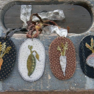 May include: Four oval-shaped felt ornaments with embroidered images of vegetables. The ornaments are black, white, brown, and dark green with white polka dots. The vegetables are a carrot, a pea pod, a turnip, and a radish.