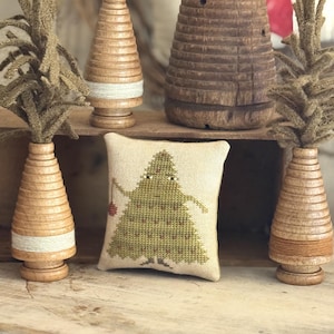 May include: A square beige fabric pincushion featuring a cross-stitched green tree with a face and a small red ornament, surrounded by four wooden spool-shaped decorative trees with textured green fiber tops and wrapped thread.