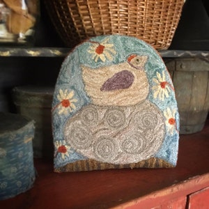 Op de afbeelding: Een blauw en bruin naaldviltkussen met een vogel die op een swirl-ontwerp zit. Het kussen is omgeven door witte madeliefjes met oranje centra.