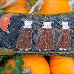 May include: A handmade needlepoint pillow with a dark blue background featuring three witches flying on a broomstick under a red crescent moon. The witches are wearing brown dresses and black hats. The pillow has a green beaded border.