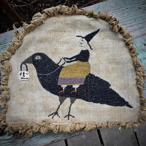 Pode incluir: Um corvo preto com uma bruxa montada em suas costas. A bruxa está usando um chapéu preto, um vestido amarelo e um cinto roxo. O corvo está segurando uma pequena bolsa branca com um rosto preto.