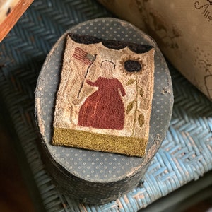 May include: A vintage needlepoint picture of a woman holding an American flag with a sunflower behind her. The picture is framed in a dark green border and is displayed on a round, black box with white polka dots.