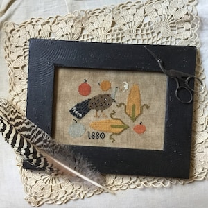Könnte beinhalten: Eine gerahmte Kreuzstich-Stickerei mit einem Vogel, Mais und Äpfeln im rustikalen Stil. Die Stickerei ist mit "1880" datiert und wird auf einer weißen Oberfläche mit einer gehäkelten Unterlage und einer Feder präsentiert.
