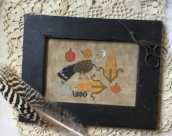 May include: A framed cross-stitch embroidery depicting a bird, corn, and apples in a rustic style. The embroidery is dated "1880" and is displayed on a white surface with a crocheted doily and a feather.