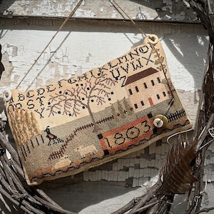 Könnte beinhalten: Ein beige-braunes Nadelfilzkissen mit einer gestickten Szene eines Hauses, Bäumen und einem Vogel. Das Kissen hat einen weißen Knopf und den gestickten Text "1803". Das Kissen hängt an einem verwitterten Holzkranz.