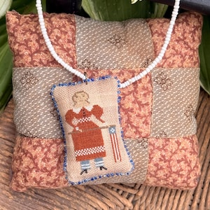 May include: A patriotic pendant necklace featuring a stitched design of a figure holding an American flag. The pendant is framed with blue beads and hangs on a white beaded chain. The pendant rests on a patchwork pillow with floral and striped patterns.