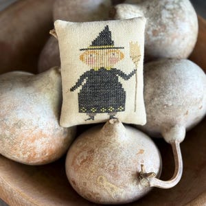 May include: A small rectangular fabric pincushion featuring a cross-stitched design of a figure wearing a black pointed hat and a black dress, holding a tan broom. The figure has orange hair, pink cheeks, and a yellow collar with matching hem details.