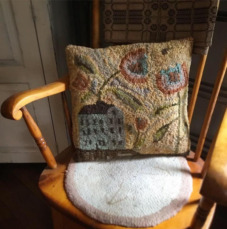 Pode incluir: Uma almofada de tapete feita &agrave; m&atilde;o com um design floral que apresenta um vaso com flores e folhas em tons de marrom, verde, azul e laranja. A almofada est&aacute; sentada em uma cadeira de balan&ccedil;o de madeira.