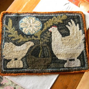 May include: A hand-stitched rug depicting two white chickens, one standing and one sitting, with a green plant in a brown pot. The rug has a brown, orange, and green color scheme and is framed with a crocheted orange border.