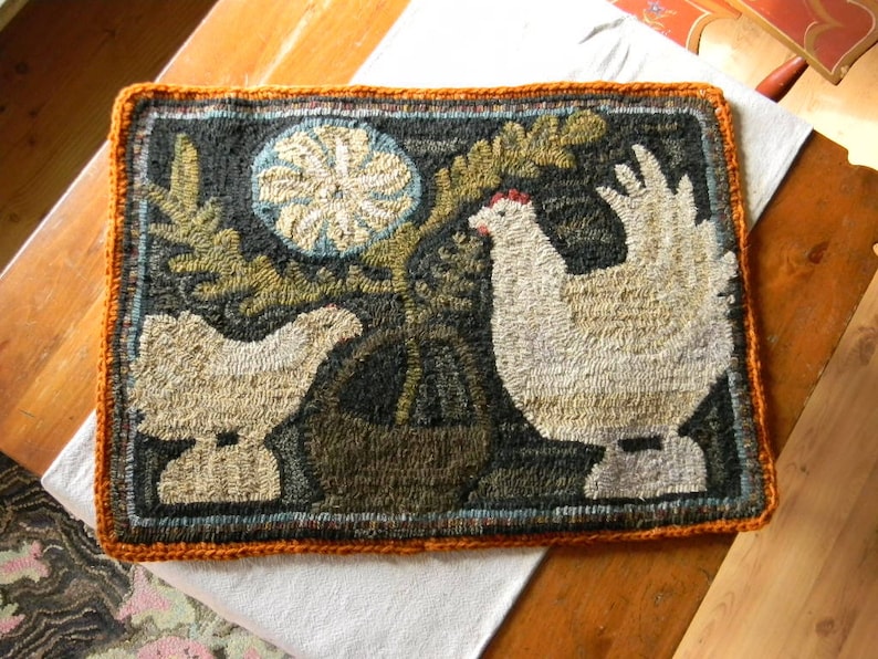 May include: A hand-stitched rug depicting two white chickens, one standing and one sitting, with a green plant in a brown pot. The rug has a brown, orange, and green color scheme and is framed with a crocheted orange border.