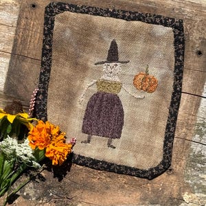 Puede incluir: Obra de arte bordada enmarcada que presenta una bruja caprichosa sosteniendo una calabaza. La bruja lleva un sombrero negro, una parte superior verde y una falda morada. La obra está enmarcada con un borde floral negro y sobre un fondo de madera rústica.