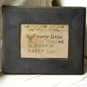 Könnte beinhalten: Ein schwarzer Holzrahmen mit weißem Hintergrund und dem Text "very little is needed to make a happy life." Der Text ist in einer dekorativen Schriftart gehalten und von einem Rand aus kleinen, stilisierten Blumen umgeben.