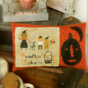 May include: A red and white pillow with a cross-stitch design featuring a group of people dressed as pumpkins. The text "Pumpkin Folke" is stitched below the figures. The pillow is sitting on a wooden surface.