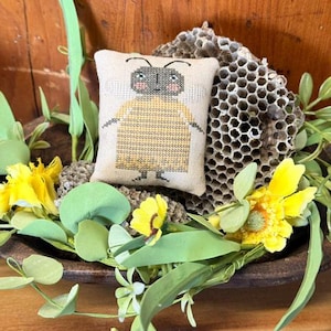 May include: A small, beige, cross-stitched bee cushion with a yellow body and grey head rests in a wooden bowl. The bowl is decorated with yellow flowers, green leaves, and a honeycomb.