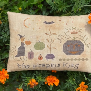 May include: A beige pillow with a cross-stitch design featuring a witch, pumpkins, a cauldron, and a bat. The text "the pumpkin king" is stitched at the bottom of the pillow.