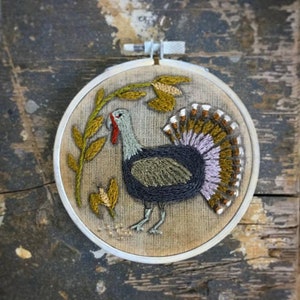 May include: Embroidered hoop art featuring a turkey with a brown, purple, and green tail feathers. The turkey is surrounded by green leaves and yellow flowers.