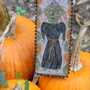 May include: A hand-stitched needlepoint wall hanging featuring a green witch with a black dress and a brown hat. The witch is surrounded by orange and green pumpkins.
