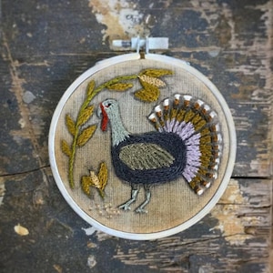 May include: Embroidered hoop art featuring a turkey with a brown, purple, and green tail feathers. The turkey is surrounded by green leaves and yellow flowers.